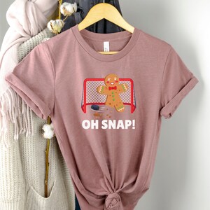 May include: A pink t-shirt with a gingerbread man playing hockey. The gingerbread man is wearing a red bow tie and is holding a hockey stick. The text "OH SNAP!" is printed on the shirt.