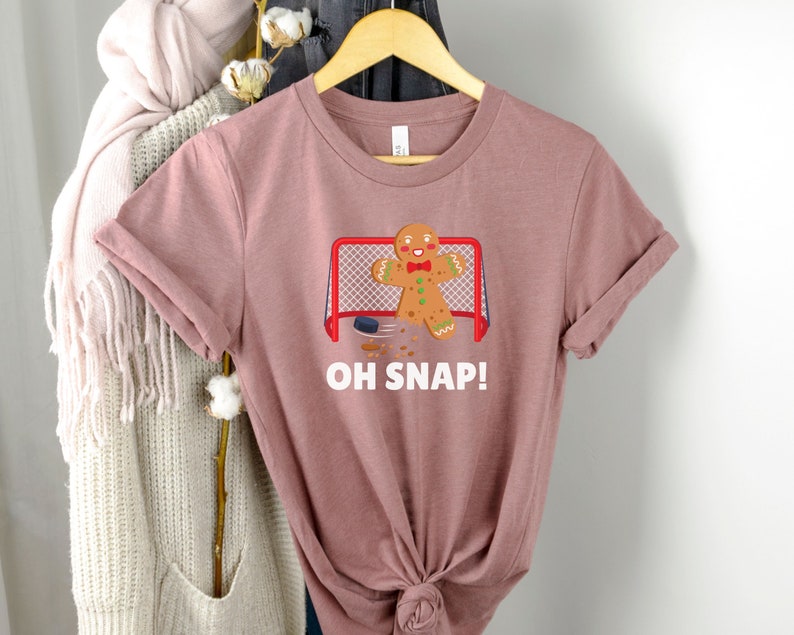 May include: A pink t-shirt with a gingerbread man playing hockey. The gingerbread man is wearing a red bow tie and is holding a hockey stick. The text "OH SNAP!" is printed on the shirt.