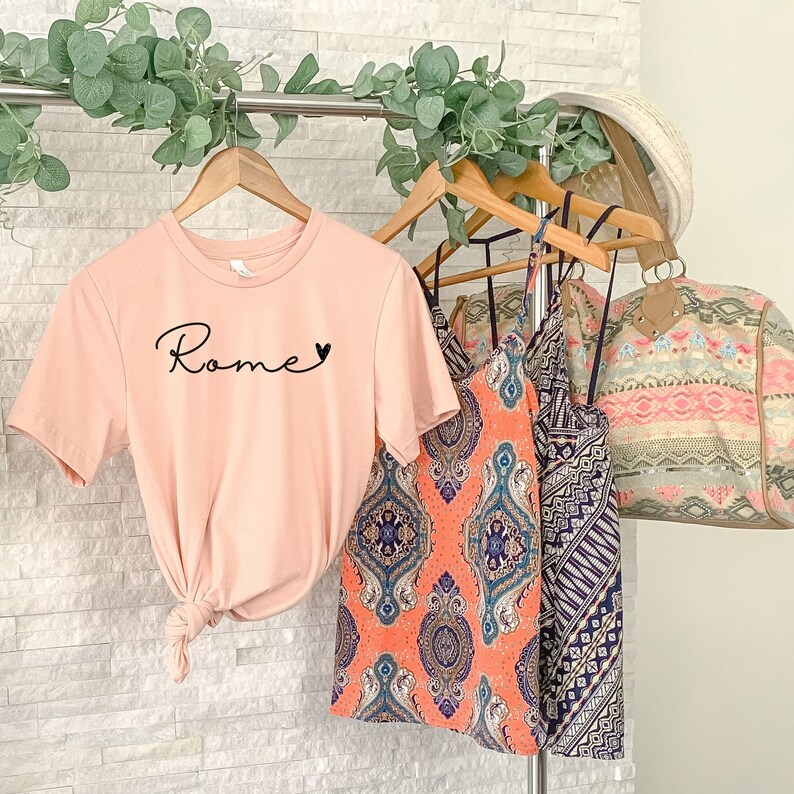 Op de afbeelding: Een roze T-shirt met het woord "Rome" en een hartprint op de voorkant. Het T-shirt hangt aan een rek met twee geprinte tanktops en een bruin en beige geprinte tas.