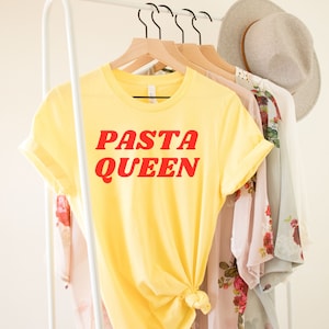 May include: Yellow t-shirt with the text "PASTA QUEEN" in red letters. The shirt is hanging on a white rack.