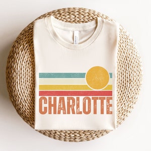 May include: A white t-shirt with a retro sunset graphic and the name "CHARLOTTE" printed in orange.