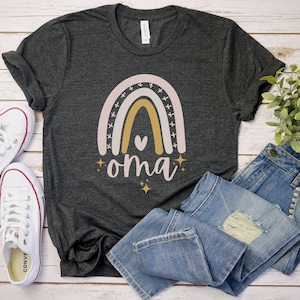 May include: A dark gray t-shirt with a rainbow design and the word "oma" in pink lettering. The rainbow has a pink, white, and gold color scheme. There are small gold stars around the rainbow and the word "oma".