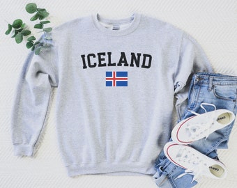 May include: Gray sweatshirt with the word "ICELAND" printed in black letters and the Icelandic flag underneath. The sweatshirt is laid flat on a white surface with a pair of blue jeans and white sneakers.
