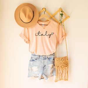 May include: A peach-colored t-shirt with the word "italy" written in black cursive script with a small Italian flag heart next to it. The shirt is hanging on a wooden hanger with a straw hat and a woven bag.