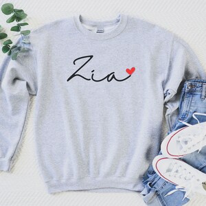 Puede incluir: Una sudadera gris con el nombre "Zia" escrito en cursiva negra con un coraz&oacute;n rojo a su lado. La sudadera est&aacute; colocada sobre una superficie blanca con un par de pantalones vaqueros azules y zapatillas blancas.
