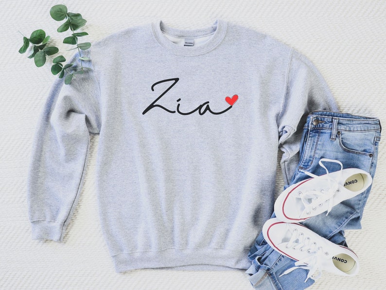 Puede incluir: Una sudadera gris con el nombre "Zia" escrito en cursiva negra con un coraz&oacute;n rojo a su lado. La sudadera est&aacute; colocada sobre una superficie blanca con un par de pantalones vaqueros azules y zapatillas blancas.