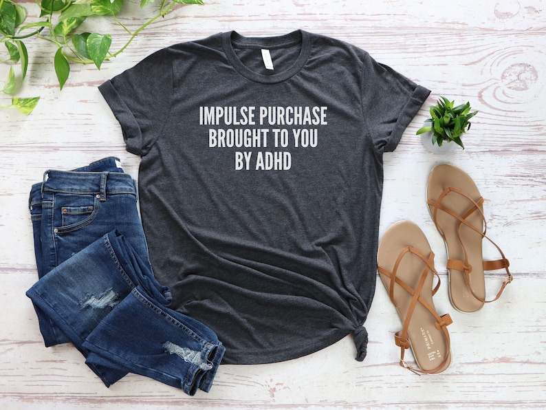 Puede incluir: Una camiseta gris oscuro con el texto "Impulse purchase brought to you by ADHD" impreso en blanco.
