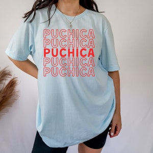 Puede incluir: Una camiseta azul claro con la palabra "Puchica" repetida cinco veces en texto rojo.