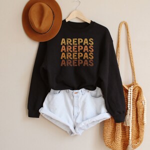 May include: Black sweatshirt with the word "AREPAS" repeated four times in a vintage style font, with each word in a different shade of orange, yellow, and brown. The sweatshirt is hanging on a wooden hanger. A brown hat and a crocheted bag are also in the image.
