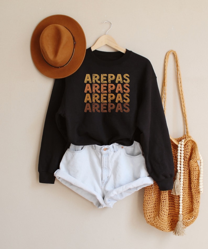 May include: Black sweatshirt with the word "AREPAS" repeated four times in a vintage style font, with each word in a different shade of orange, yellow, and brown. The sweatshirt is hanging on a wooden hanger. A brown hat and a crocheted bag are also in the image.