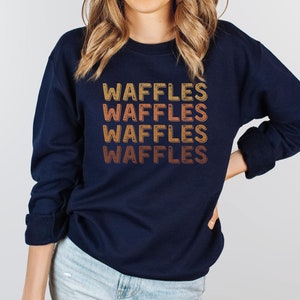 May include: A navy blue sweatshirt with the word "WAFFLES" repeated five times in a retro, gradient font. The text is in shades of orange, yellow, and brown.