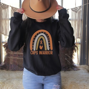 May include: Black sweatshirt with a rainbow design and the text "CRPS Warrior". The rainbow is made up of orange, yellow, and white stripes with a brown ribbon and a heart in the center.