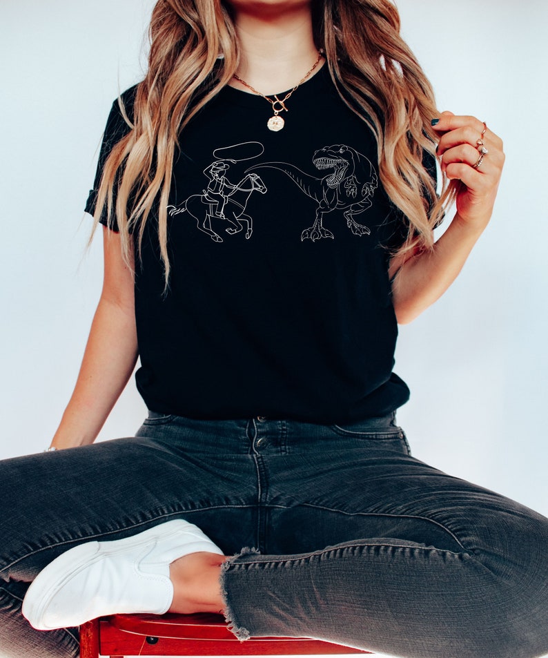 May include: A black t-shirt with a white graphic of a cowboy riding a horse and lassoing a dinosaur.