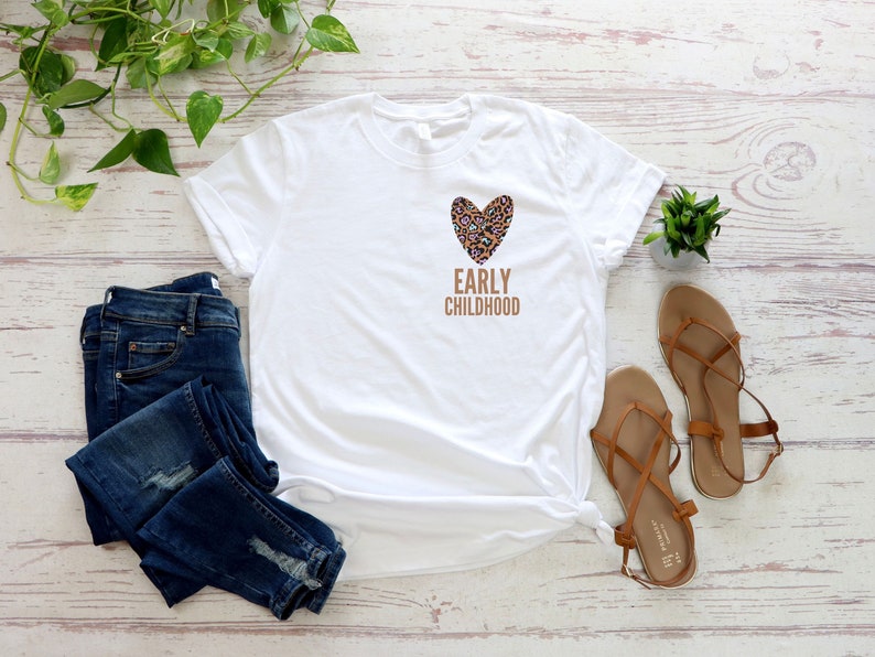 May include: White t-shirt with a brown leopard print heart and the text "EARLY CHILDHOOD" printed on the front. The shirt is paired with blue jeans and brown sandals.