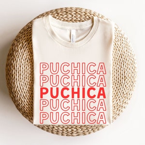 Puede incluir: Una camiseta blanca con la palabra "Puchica" repetida cinco veces en rojo, con contorno blanco. La camiseta est&aacute; doblada y colocada sobre una estera de paja tejida.