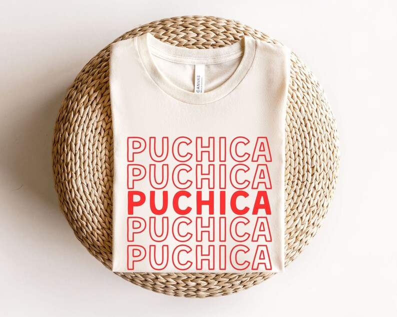 Puede incluir: Una camiseta blanca con la palabra "Puchica" repetida cinco veces en rojo, con contorno blanco. La camiseta est&aacute; doblada y colocada sobre una estera de paja tejida.
