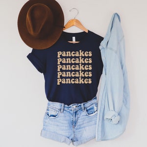 Pode incluir: Uma camiseta azul marinho com a palavra "Pancakes" repetida cinco vezes em uma fonte retr&ocirc; de cor marrom claro. A camiseta est&aacute; pendurada em um cabide de madeira e &eacute; combinada com um chap&eacute;u fedora marrom, shorts jeans azul e uma jaqueta jeans azul claro.