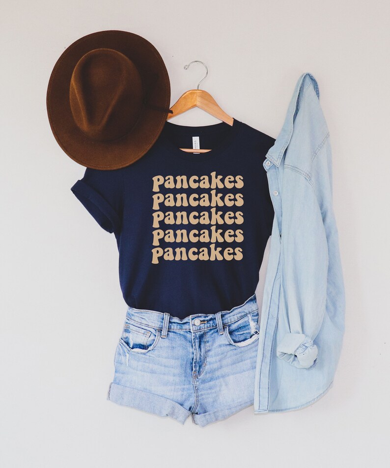 Pode incluir: Uma camiseta azul marinho com a palavra "Pancakes" repetida cinco vezes em uma fonte retr&ocirc; de cor marrom claro. A camiseta est&aacute; pendurada em um cabide de madeira e &eacute; combinada com um chap&eacute;u fedora marrom, shorts jeans azul e uma jaqueta jeans azul claro.