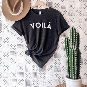 May include: A dark grey heather t-shirt with the word "VOILA" printed in white on the front. The shirt is hanging on a wooden hanger against a white wall with a black and white geometric pattern.