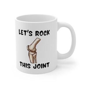 May include: White ceramic coffee mug with a black graphic of a knee joint and the text "Let's Rock This Joint".