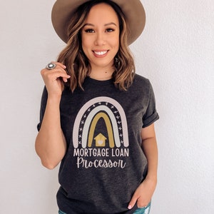 May include: Dark gray t-shirt with a rainbow design and the words "MORTGAGE LOAN PROCESSOR" in a script font. The shirt features a house graphic within the rainbow. The person is wearing a brown hat and blue jeans.