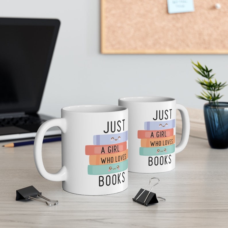 Op de afbeelding: Twee witte keramische mokken met een zwart-witte afbeelding van een stapel boeken met glimlachende gezichten. De tekst op de mokken luidt "Just a girl who loves books".
