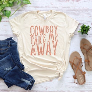 May include: A beige t-shirt with the text "COWBOY TAKE ME AWAY" printed in a pink color on the front.