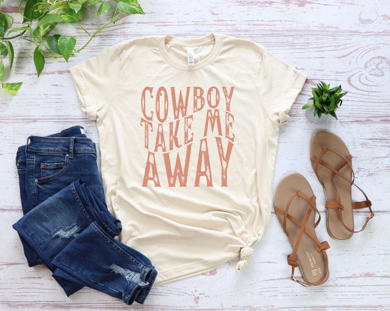 May include: A beige t-shirt with the text "COWBOY TAKE ME AWAY" printed in a pink color on the front.