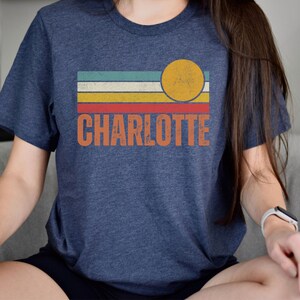 May include: A navy blue t-shirt with a vintage-style design featuring a yellow sun and colorful stripes. The text "CHARLOTTE" is printed in orange below the design.