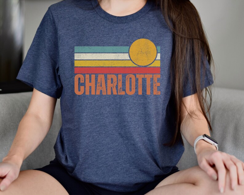 May include: A navy blue t-shirt with a vintage-style design featuring a yellow sun and colorful stripes. The text "CHARLOTTE" is printed in orange below the design.