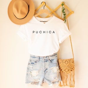 May include: White t-shirt with the word "Puchica" printed in black on the front. The t-shirt is hanging on a wooden hanger with a straw hat and a crocheted bag.