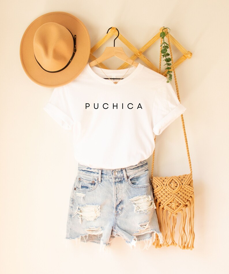 May include: White t-shirt with the word "Puchica" printed in black on the front. The t-shirt is hanging on a wooden hanger with a straw hat and a crocheted bag.