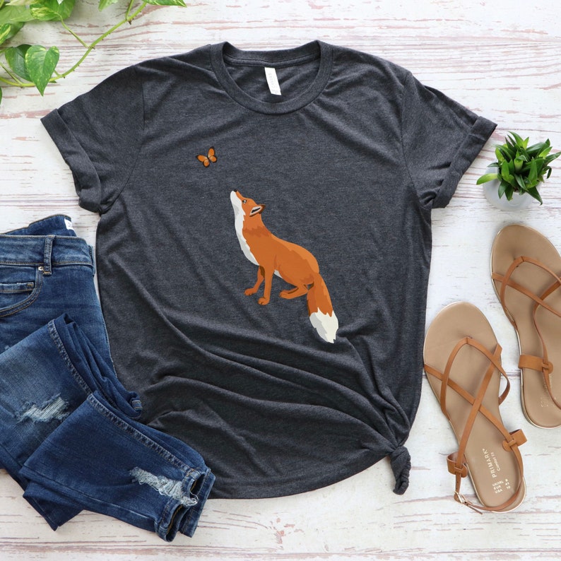 Op de afbeelding: Een donkergrijs T-shirt met een afbeelding van een vos die naar een vlinder kijkt. De vos is oranje en wit, en de vlinder is oranje.