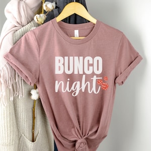 May include: A mauve t-shirt with the text "BUNCO night" in white lettering and red dice graphics.