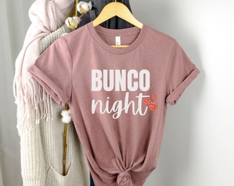 May include: A mauve t-shirt with the text "BUNCO night" in white lettering and red dice graphics.