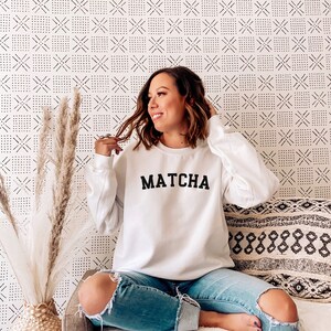 May include: A white sweatshirt with the word "MATCHA" printed in black on the front. The sweatshirt is being worn by a person sitting on a white couch with a patterned throw pillow.