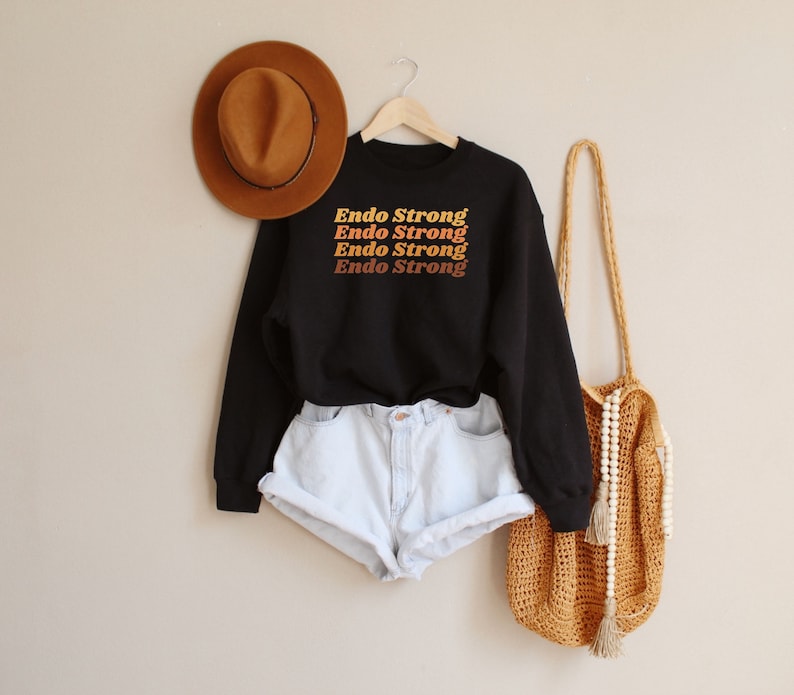 May include: Black sweatshirt with the text "Endo Strong" repeated four times in a gradient of orange, yellow, and red. The sweatshirt is worn with white denim shorts and a brown woven tote bag with white beads.