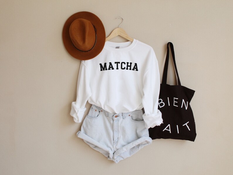 May include: White sweatshirt with the word "MATCHA" printed in black. The sweatshirt is paired with light blue denim shorts and a black tote bag with the words "BIEN FAIT" printed in white.