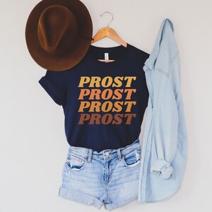 May include: A navy blue t-shirt with the word "PROST" repeated four times in a retro font, with each word in a different shade of orange, brown, and yellow. The shirt is hanging on a wooden hanger and is styled with a brown hat and a light blue denim jacket.