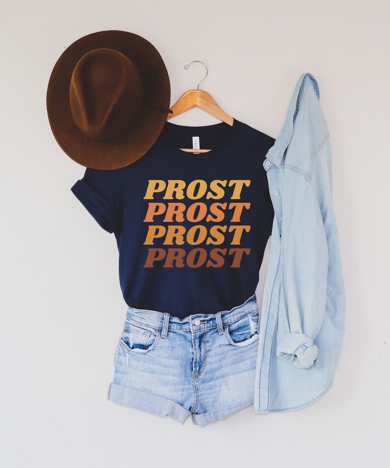 May include: A navy blue t-shirt with the word "PROST" repeated four times in a retro font, with each word in a different shade of orange, brown, and yellow. The shirt is hanging on a wooden hanger and is styled with a brown hat and a light blue denim jacket.