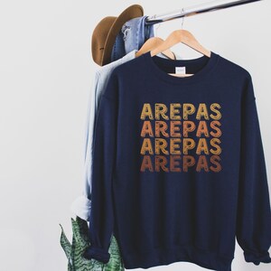 May include: A navy blue sweatshirt with the word "AREPAS" repeated four times in a vintage style font. The text is in shades of orange, yellow, and red.
