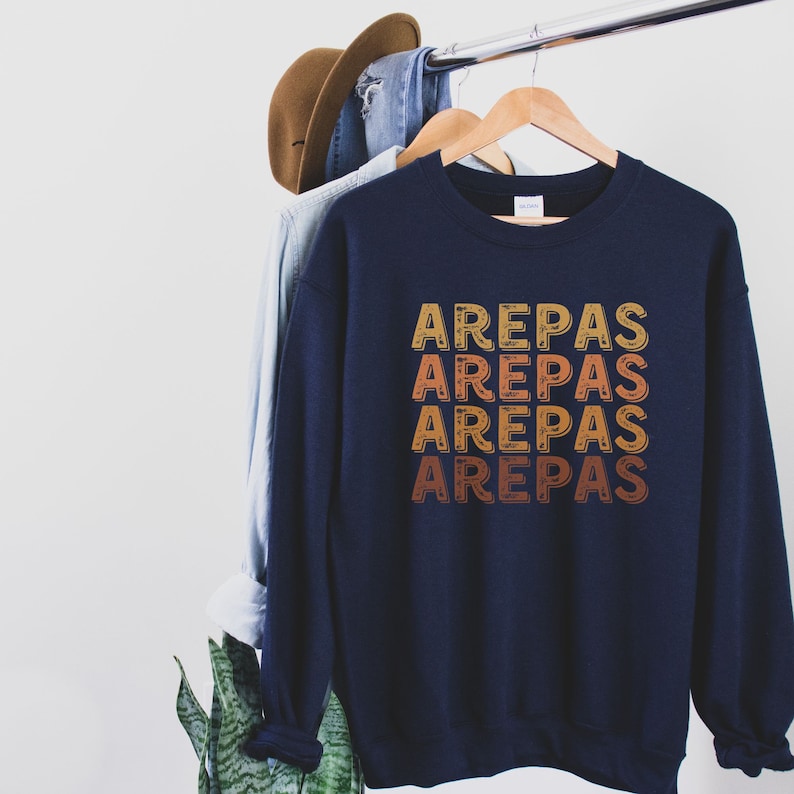 May include: A navy blue sweatshirt with the word "AREPAS" repeated four times in a vintage style font. The text is in shades of orange, yellow, and red.
