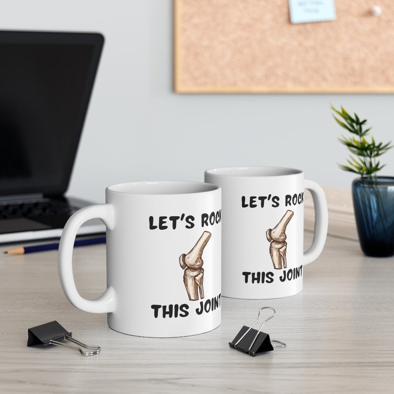 May include: Two white ceramic mugs with black text and a cartoon knee bone. The text on the mugs reads "Let's Rock This Joint".