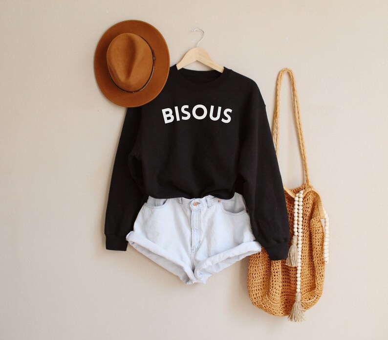 May include: A black sweatshirt with the word "BISOUS" in white lettering. The sweatshirt is paired with light blue denim shorts, a brown fedora hat, and a woven shoulder bag.