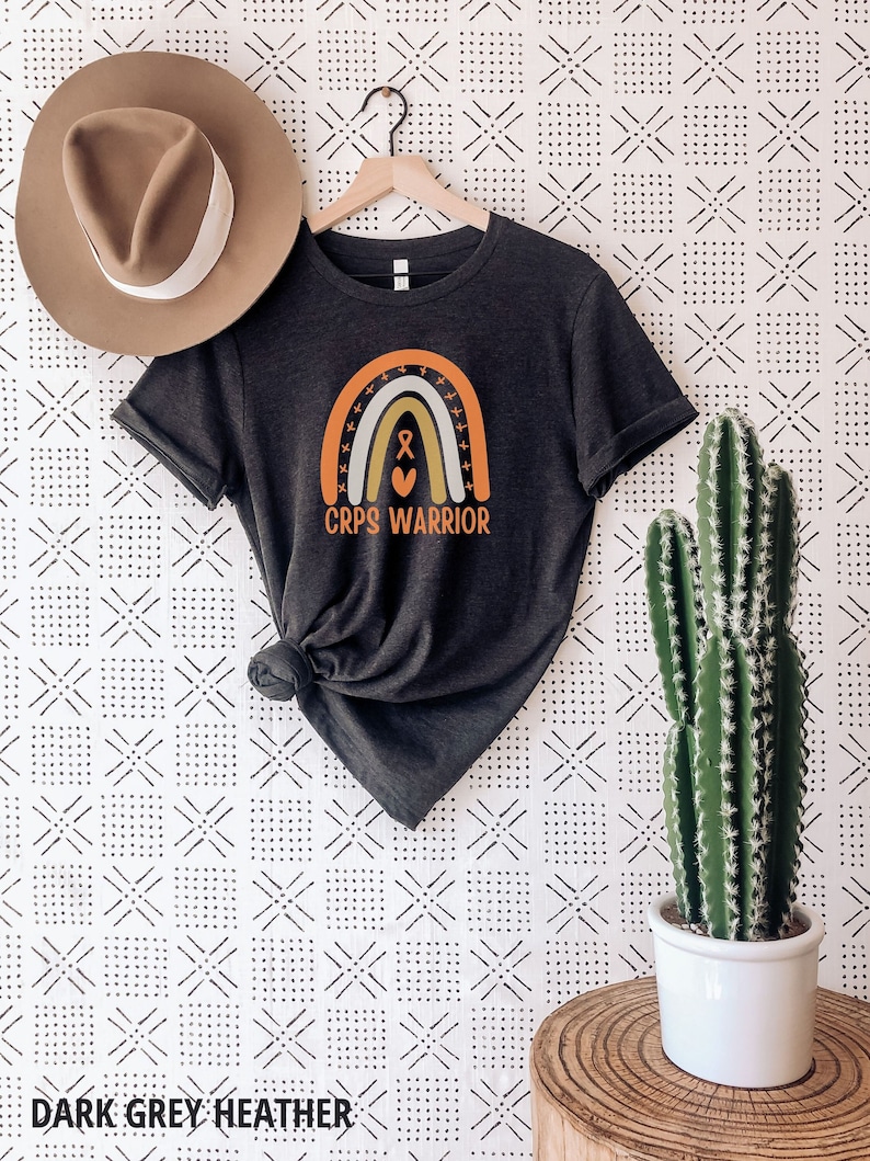 May include: A dark grey heather t-shirt featuring a rainbow graphic and the text "CRPS WARRIOR". The rainbow has a heart and an orange ribbon in the centre.