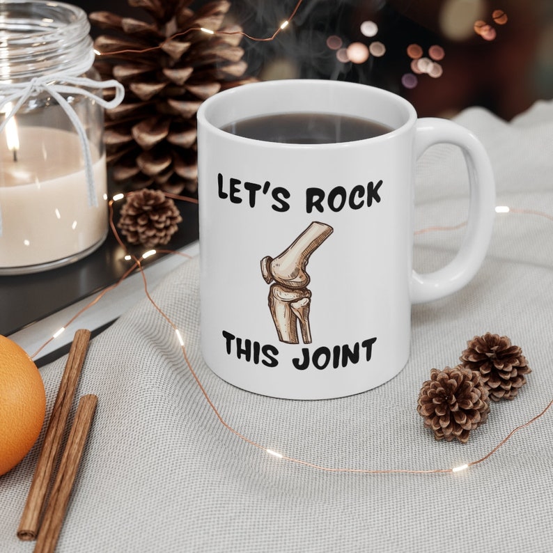 May include: White ceramic coffee mug with a black graphic of a knee bone and the text "Let's Rock This Joint".