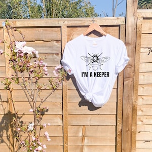 May include: White t-shirt with a black bee graphic and the text "I'm a Keeper".
