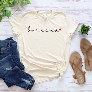 May include: Cream-colored t-shirt with the word "boricua" in black script and a small red heart. The shirt is paired with blue jeans and brown sandals. A small green plant is also visible.