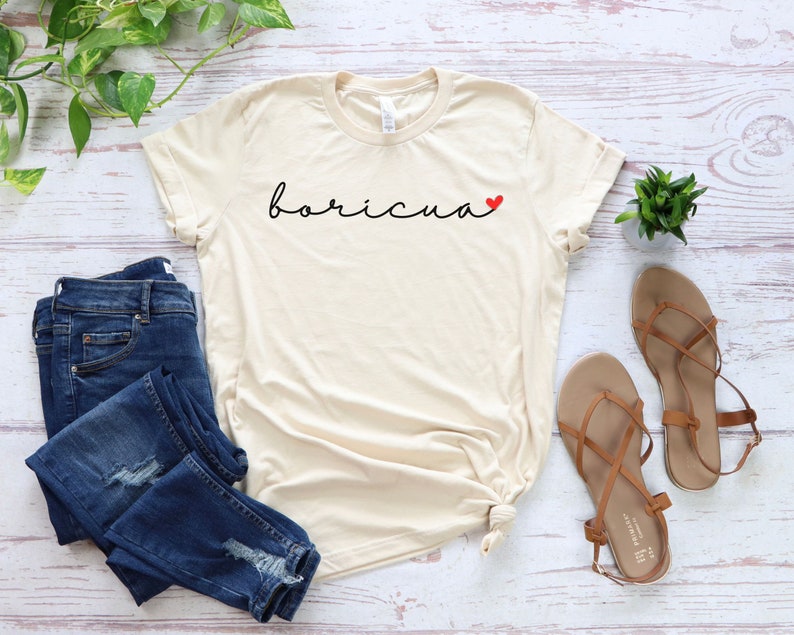 May include: Cream-colored t-shirt with the word "boricua" in black script and a small red heart. The shirt is paired with blue jeans and brown sandals. A small green plant is also visible.