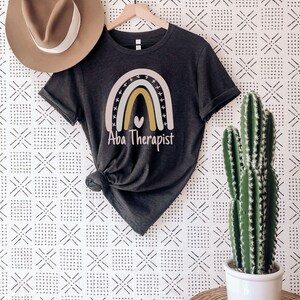 May include: A dark grey heather t-shirt with a rainbow graphic and the text "Aba Therapist".